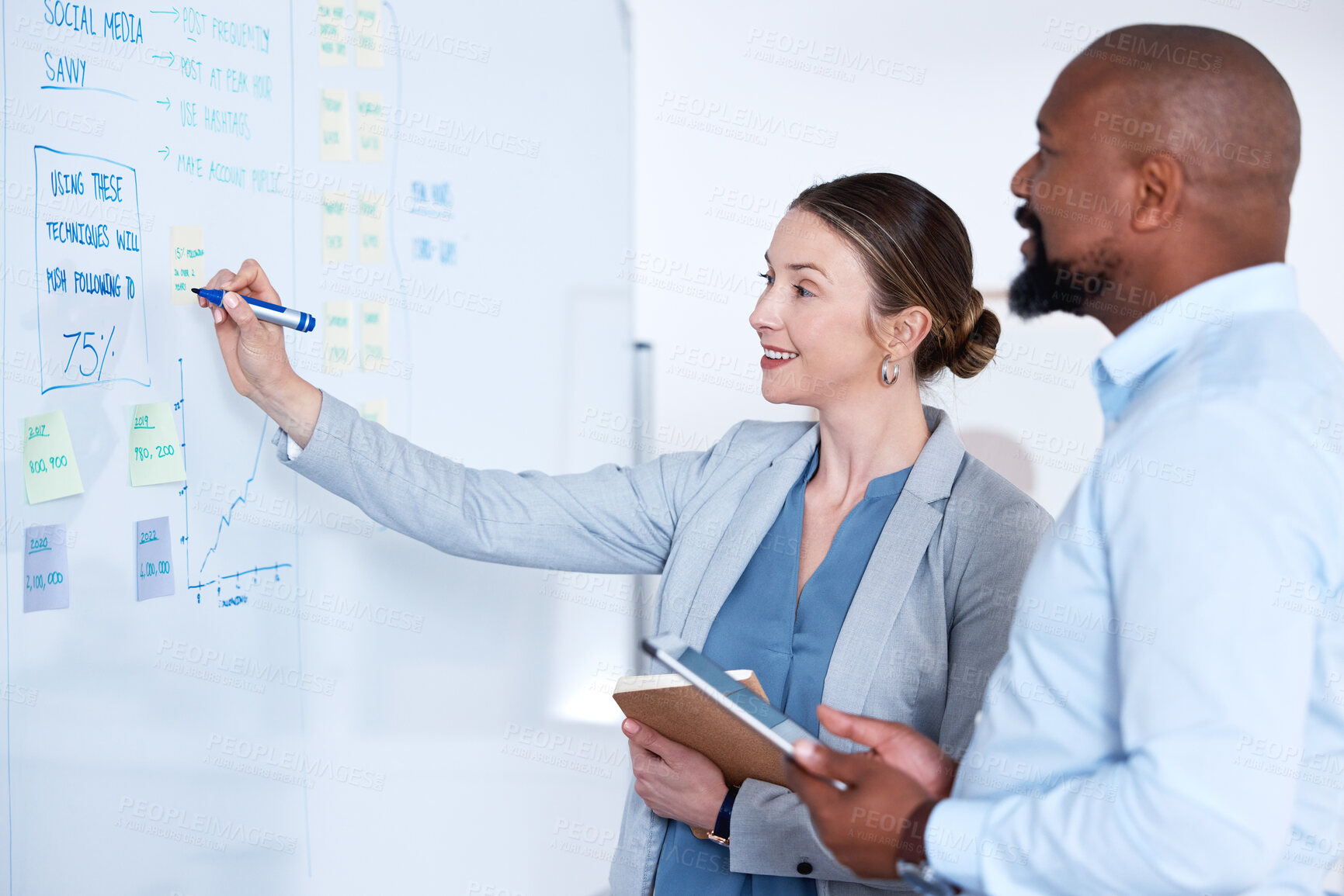 Buy stock photo Team of smiling business people using whiteboard to brainstorm strategy in office. Caucasian businesswoman standing with african american businessman and writing while planning. Two happy colleagues