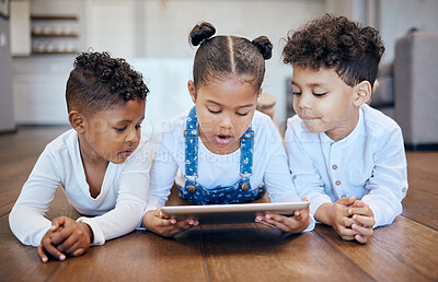 Three young adorable mixed race children using a digital tablet while lying together on the wooden floor at home. Two young boys and girl siblings browsing the internet online to play games and learn from educational apps