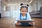 One adorable little mixed race girl lying down on a home living room floor and watching education videos on a digital tablet. Cute hispanic child listening to music with headphones and playing games