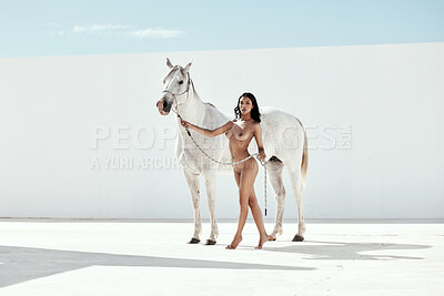 Full Body of beautiful mixed race woman posing nude while standing next to a white horse outdoors. Sexy hot hispanic model showing her naked body with white stallion. Nudist feeling sensual, seductive, free on a mare