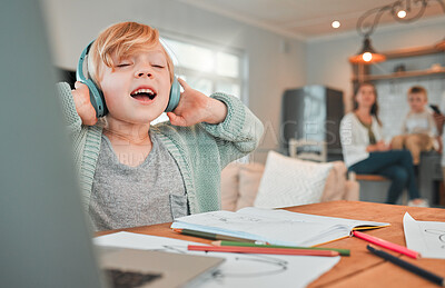 A cute caucasian little blonde girl wearing wireless headphones while being creative by drawing in a book at home with her family in the background. Girl enjoying magic of music ignoring homework