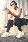 One fit young hispanic woman listening to music with headphones from a cellphone while taking a break from exercise outdoors. Female athlete texting and using fitness apps online while browsing social media and watching workout tutorials