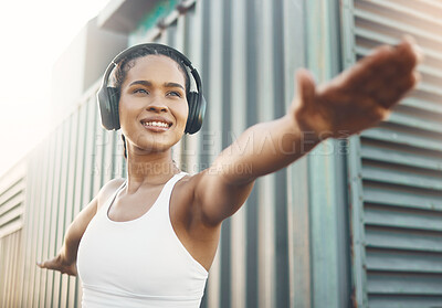 One fit young hispanic woman stretching arms in warrior pose for warmup to prevent injury while exercising in an urban setting outdoors. Happy and motivated female athlete listening to music with headphones while preparing body and mind for training