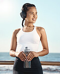 Happy young hispanic female athlete smiling while listening to music on headphones and holding water bottle while taking a break during her cardio workout exercise along the seaside promenade