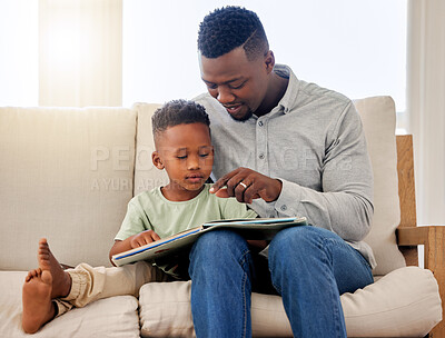 African american dad pointing at picture in storybook. Adorable little boy sitting with his father and reading a storybook together on the couch at home . Father and son spending time together