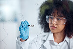 One serious young mixed race female scientist with a curly afro writing and planning equations on a glass board wearing glasses and gloves at work. Focused hispanic lab worker drawing on a board looking determined