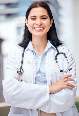 Confident young female doctor standing with her arms crossed outside a medical office. One caucasian woman in a white coat with stethoscope. Trusted practitioner caring for the health of patients