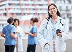 Portrait of smiling young doctor enjoying a cup of coffee outside with her colleagues in the background. Happy medical professional taking a break from working in the hospital to drink tea. 
