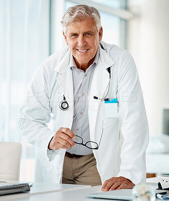 Buy stock photo Experienced mature caucasian man doctor smiling and standing and holding his glasses in a hospital. One optimistic senior male general practitioner wearing a lab coat looking confident and happy to give patient good news while leaning on a desk