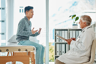A young asian man talking while consulting with a caucasian senior doctor while sitting on a bed in the clinic. Mixed race patient explaining as he receives advice from his doctor about his medical health and wellness at a hospital. Doctor taking notes