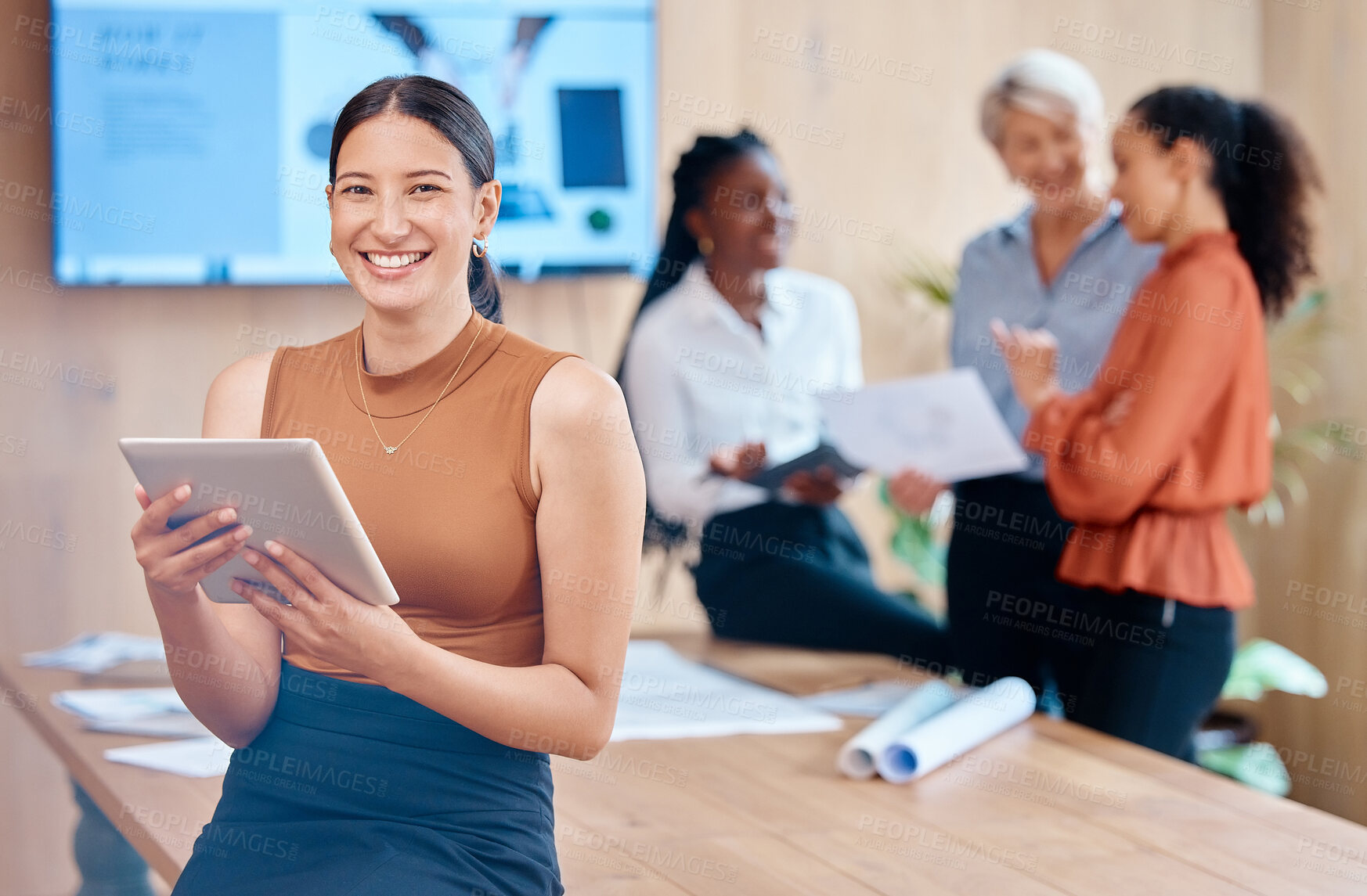 Buy stock photo Beautiful young mixed race business woman using a tablet while leaning against the boardroom table after a meeting with her female only colleagues in the background. Our office is going digital