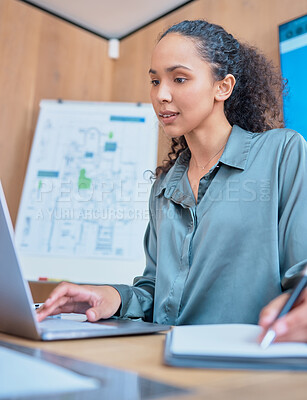 Young happy mixed race businesswoman with curly hair writing in a notebook and working on a laptop sitting at a desk in an office at work. One hispanic female writing a note and typing an email