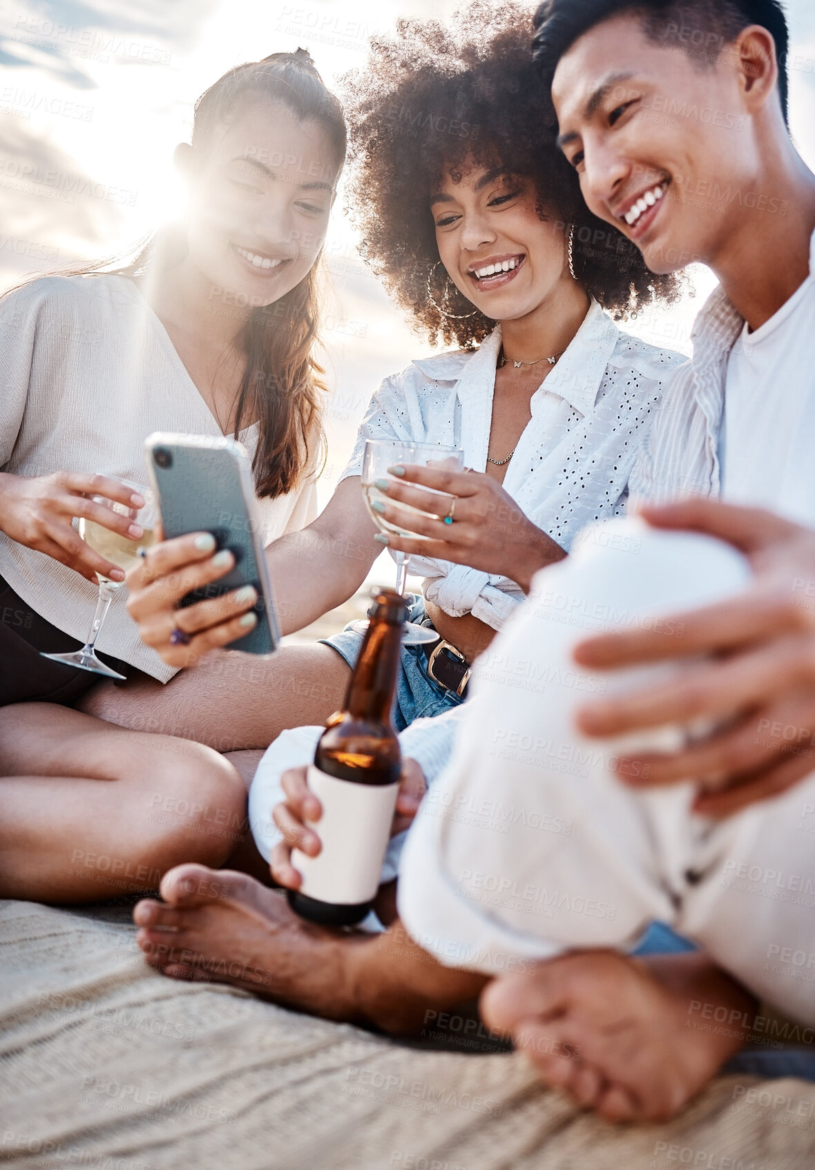 Buy stock photo A group three  multiracial friends relaxing and using a phone at the beach while having alcoholic drinks