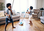 Two mixed race little boys playing with a mop and broom in the lounge at home. Happy siblings having fun playing with cleaning supplies at home. Carefree brothers singing while cleaning at home