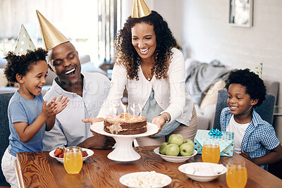 African american family celebrating birthday at home. Mom putting birthday cake with candles on table. . Two little boys and their parents wearing party hats and looking excited
