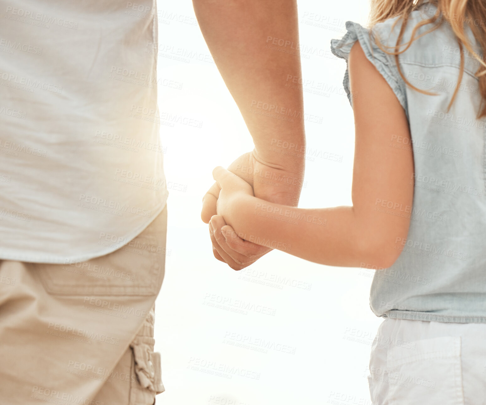 Buy stock photo Rear view of a Unrecognizable caucasian man holding the hand of his little girl while standing outdoors on a sunny day with a flare in between them. Father and young daughter showing affection and love by holding hands, a symbol of a loving family who are there to support one another