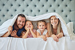 A happy family in bed under a blanket at home. Portrait of  smiling young parents having fun with children in the bedroom, covering with a duvet. Cute little girls playing with their mother and father