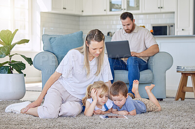 Family enjoying a movie together. A happy young family looking at a tablet together while lying on the carpet in their lounge while their father uses a laptop