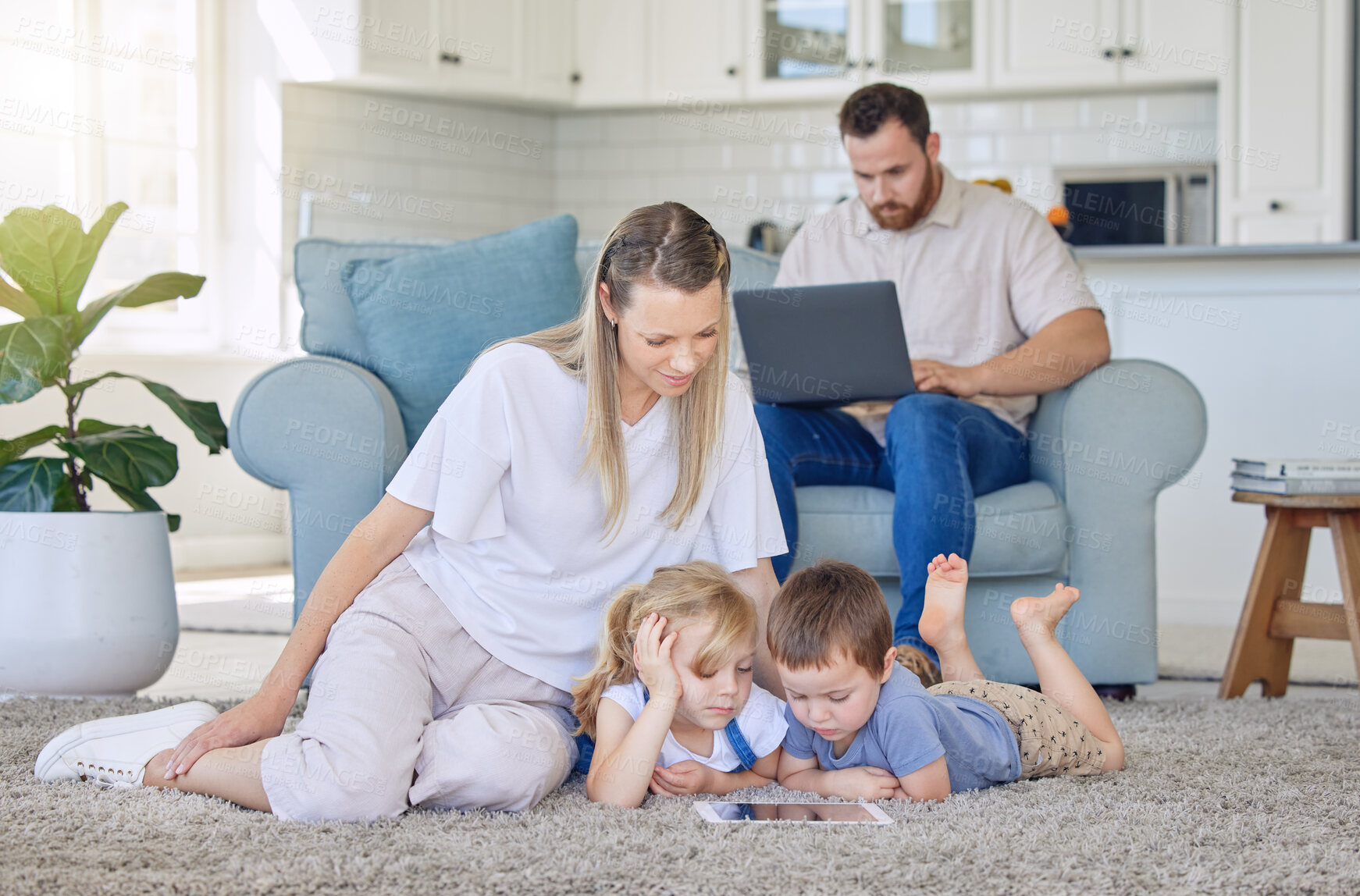 Buy stock photo Family enjoying a movie together. A happy young family looking at a tablet together while lying on the carpet in their lounge while their father uses a laptop
