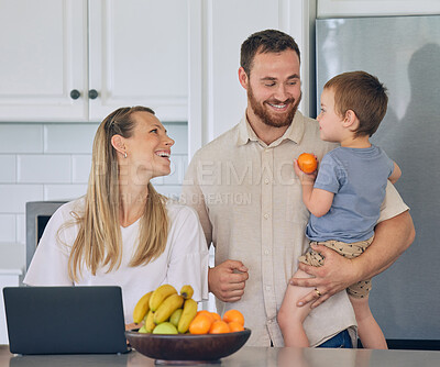 Happy young caucasian family having lunch time with fresh fruit in bright kitchen. Beautiful young mother and father feeding their cute son an orange while using a laptop at home