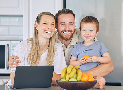 Happy family relaxing in the kitchen. Little boy eating orange in the kitchen. Cheerful caucasian family at home. Father hugging wife and son. Two parents bonding with their son. Young family laughing while mother uses a laptop