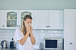 Sick caucasian woman holding tissue and blowing her nose while standing in kitchen at home. Woman suffering from seasonal allergy or flu