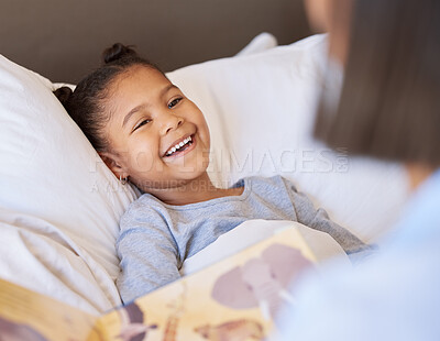 A cute little smiling mixed race girl laughing while lying in bed and enjoying story time. Young mother reading her adorable african American daughter a bedtime story while she relax in bed at home