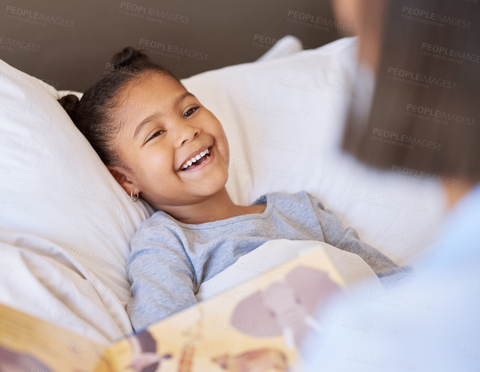 Buy stock photo A cute little smiling mixed race girl laughing while lying in bed and enjoying story time. Young mother reading her adorable african American daughter a bedtime story while she relax in bed at home 