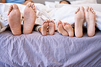 Feet of family lying in bed. Closeup of feet of parents and children side by side in bed. Family relaxing in bed together. Below bare feet of family in bed. Kids resting in bed with their parents.