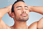 Close up of muscular asian man showering alone and washing his hair against a blue studio background. Fit and strong mixed race man standing under pouring water. Hispanic athlete enjoying hot shower with his eyes closed