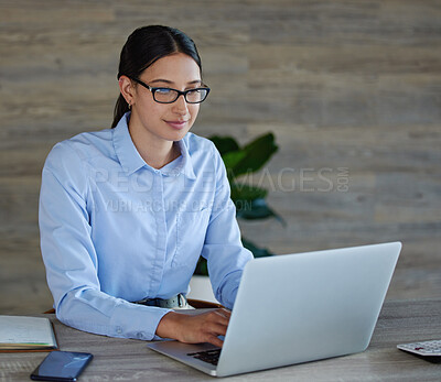 Young mixed race businesswoman wearing glasses sitting and working on a laptop alone in an office at work. One happy hispanic woman typing an email on a laptop working at desk at work