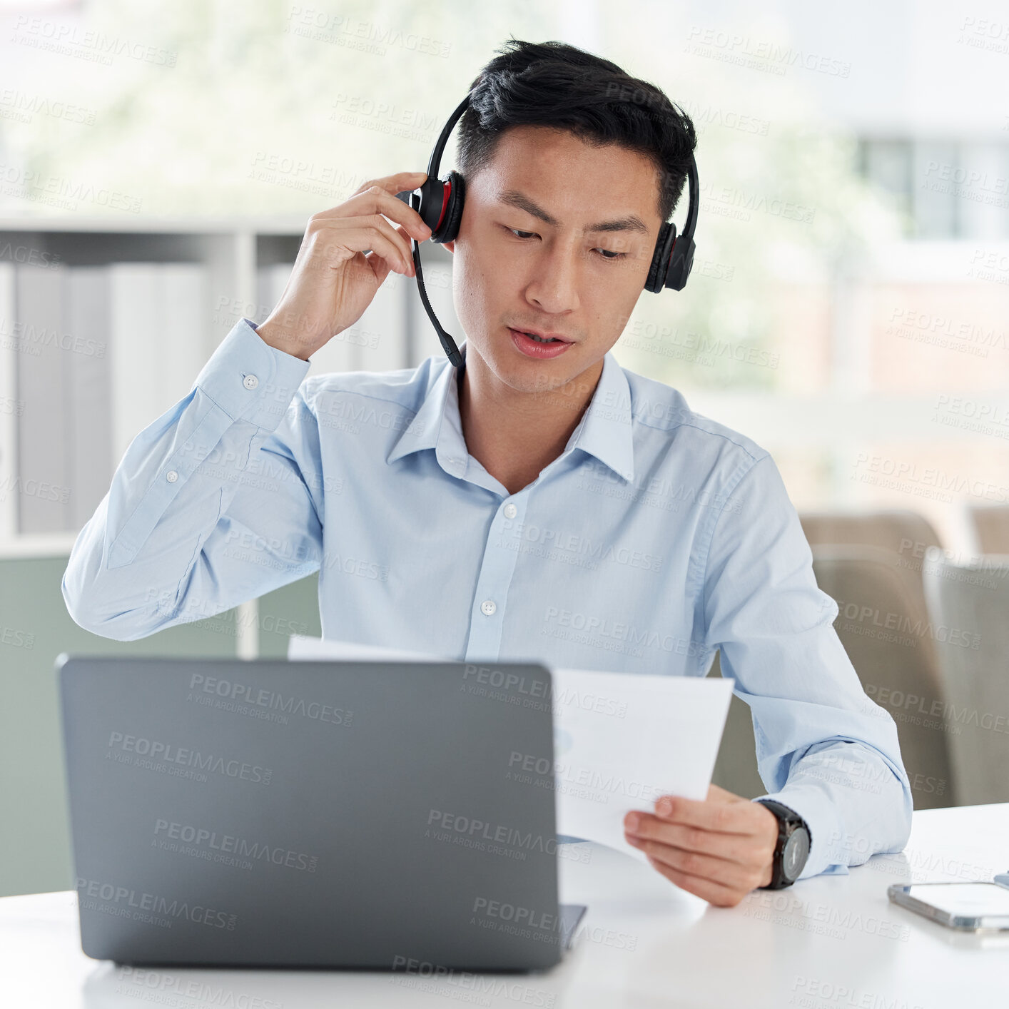 Buy stock photo One young asian call centre agent talking on headset while reading document with laptop in office. Salesman and marketing rep consultant advising while operating helpdesk for customer service support