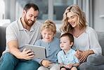 Happy relaxed family of four using a digital tablet device while bonding on the sofa together at home. Young couple checking social media and watching a movie with their two cute caucasian sons 