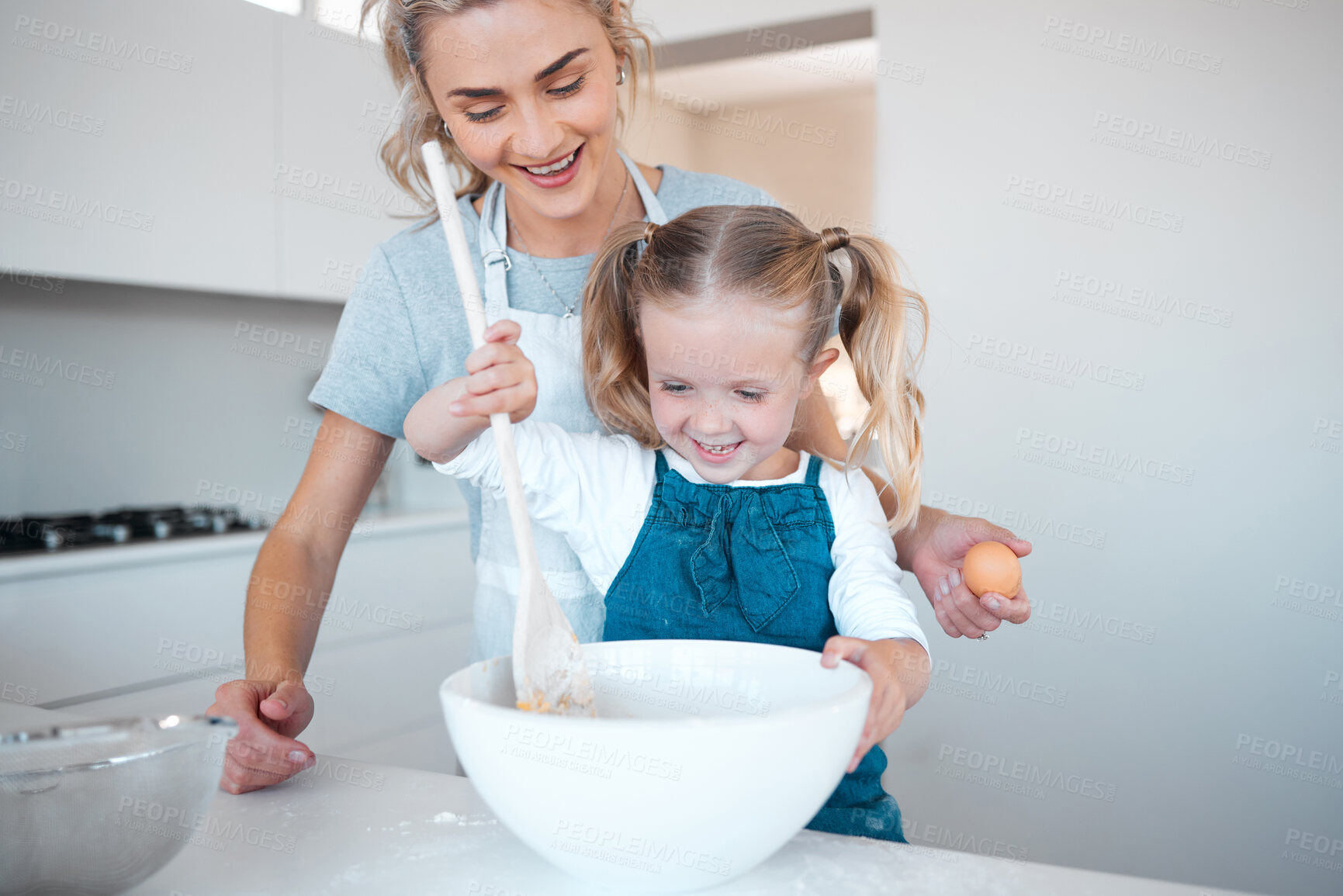 Buy stock photo Happy mother and daughter baking and bonding. Young woman helping her daughter bake at home. Smiling mother holding an egg, cooking with her daughter. Happy little mixing a bowl of batter