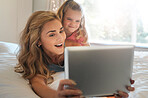 Happy young caucasian mother and daughter using a digital tablet together relaxing on a bed at home. Cute little girl learning to use a digital tablet with her mom
