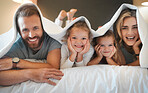 Portrait of a happy caucasian family lying in bed together under a blanket at home. Cheerful siblings relaxing in bed with their mom and dad in a bedroom