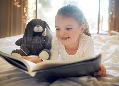 Happy smiling little girl lying on a bed at home and reading a children\'s book to her toy teddy. Cute child relaxing and playfully reading a story to her bunny, being creative and playing pretend