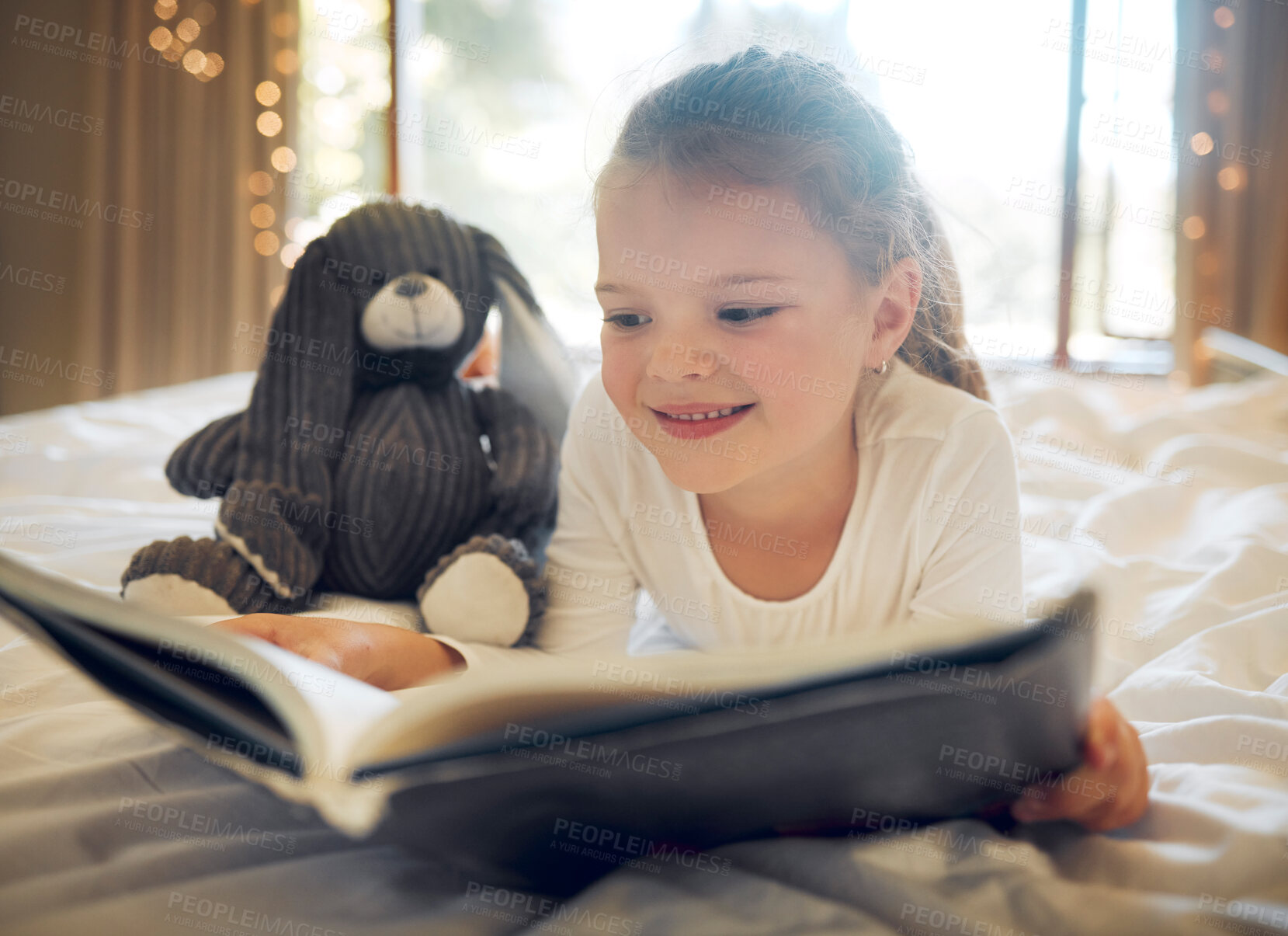 Buy stock photo Happy smiling little girl lying on a bed at home and reading a children's book to her toy teddy. Cute child relaxing and playfully reading a story to her bunny, being creative and playing pretend