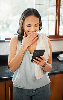 A woman using a phone and laughing at home. Happy young woman reading text message or chatting on social media while busy with chores in the kitchen