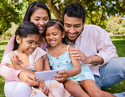 Young happy mixed race family using a phone and sitting on grass together in a park. Loving hispanic parents watching videos with their little daughters on a cellphone in nature