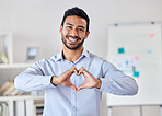 Young happy mixed race businessman making a heart gesture with his hands standing in an office alone at work. One hispanic male boss smiling showing love and support with a heart hand gesture