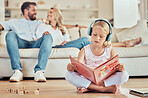 Little caucasian girl reading a story book while wearing headphones listening to music with her parents in the background. Child reading a fairytale and listening to music with her mom and dad at home