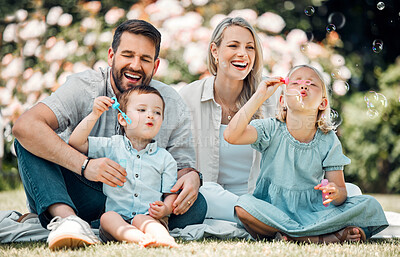 Buy stock photo Smiling caucasian family blowing soap bubbles for fun while relaxing together in the park or garden outside on a sunny day. Loving parents bonding with playful kids enjoying a carefree happy childhood