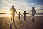 Rear view shot of a carefree family holding hands while walking into the sunset. Mixed race parents and their two kids spending time together by the sea enjoying summer vacation. Family beach day