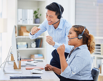 Two happy young diverse call centre telemarketing agent cheering with joy and punching the air with fists while working in an office. Excited african american and asian assistants celebrating successful sales and reaching targets to win