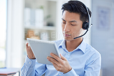One young asian male call centre telemarketing agent talking on a headset while working on a digital tablet device in an office. Focused businessman consultant browsing internet apps and interacting with clients online. Supporting customer service sales