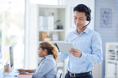 One young asian male call centre telemarketing agent talking on a headset while working on a digital tablet device in an office. Smiling businessman consultant browsing internet apps and interacting with clients online. Supporting customer service sales