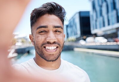Young mixed race sportsman smiling and holding mobile phone with hand to take a selfie while out for a run or jog along a water canal in the city. Happy male athlete taking self-portrait during workout