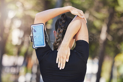 Rear view of fit young female athlete stretching her hands behind her back while listening to music on smart phone in armband. Sportswoman warming up before cardio workout at park