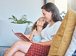Adorable little daughter looking up at her mother as she reads to her while sitting together. Mother and little girl enjoying interesting storybook about fairytales at home